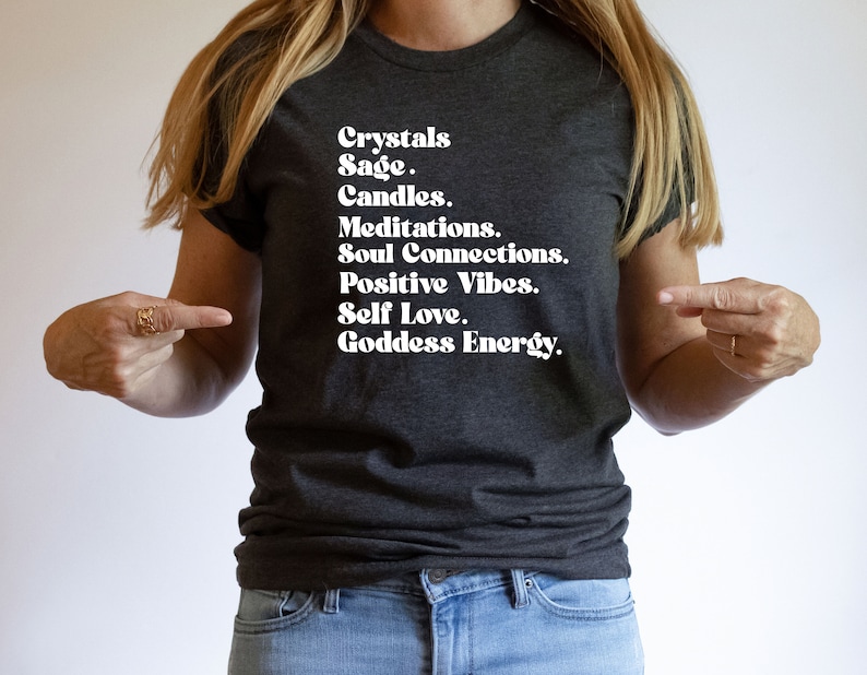 May include: A dark grey t-shirt with white text that reads "Crystals Sage. Candles. Meditations. Soul Connections. Positive Vibes. Self Love. Goddess Energy."