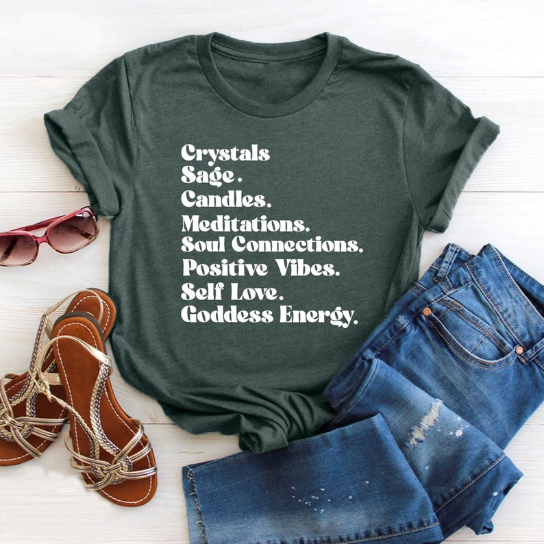 May include: A dark green t-shirt with white text that reads "Crystals Sage. Candles. Meditations. Soul Connections. Positive Vibes. Self Love. Goddess Energy."