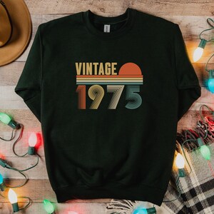 Pode incluir: Camisola verde escuro com o texto "VINTAGE 1975" num design retro. O texto &eacute; em laranja, amarelo e verde-azulado. A camisola est&aacute; numa superf&iacute;cie de madeira com luzes de Natal e um chap&eacute;u.