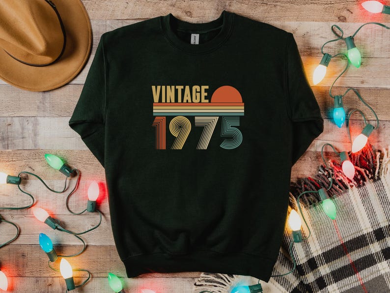 Pode incluir: Camisola verde escuro com o texto "VINTAGE 1975" num design retro. O texto &eacute; em laranja, amarelo e verde-azulado. A camisola est&aacute; numa superf&iacute;cie de madeira com luzes de Natal e um chap&eacute;u.