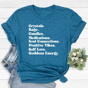May include: Teal blue t-shirt with white text that reads "Crystals, Sage, Candles, Meditations, Soul Connections, Positive Vibes, Self Love, Goddess Energy."
