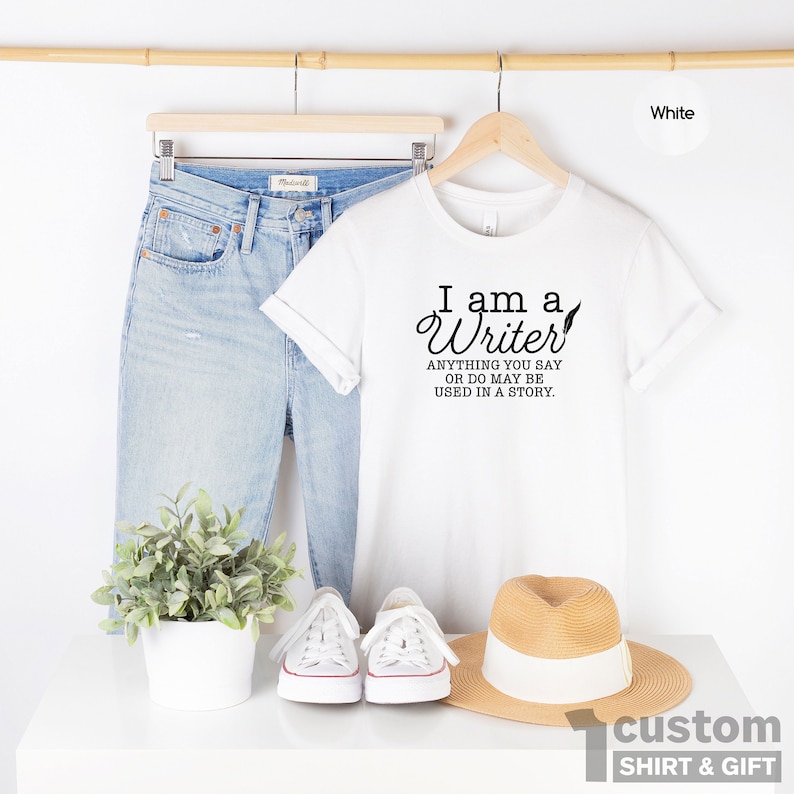 May include: White t-shirt with the text "I am a Writer. Anything you say or do may be used in a story."  The shirt is displayed on a hanger with a pair of blue jeans, a hat, and a plant.
