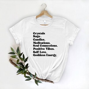 May include: White t-shirt with black text that reads "Crystals, Sage, Candles, Meditations, Soul Connections, Positive Vibes, Self Love, Goddess Energy."