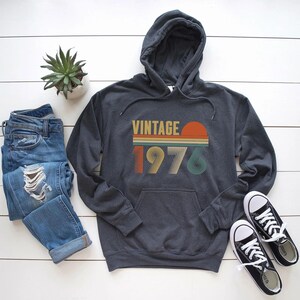 Pode incluir: Moletom cinza escuro com um design retr&ocirc; "VINTAGE 1976" em laranja, amarelo e verde. O moletom tem um bolso frontal e um capuz. A imagem tamb&eacute;m inclui um par de jeans azuis, t&ecirc;nis pretos e uma pequena planta em vaso.