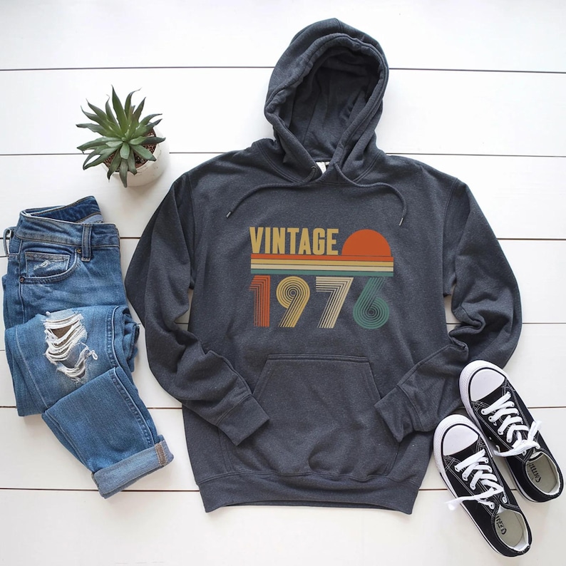 Pode incluir: Moletom cinza escuro com um design retr&ocirc; "VINTAGE 1976" em laranja, amarelo e verde. O moletom tem um bolso frontal e um capuz. A imagem tamb&eacute;m inclui um par de jeans azuis, t&ecirc;nis pretos e uma pequena planta em vaso.