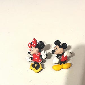 May include: Two cartoon figurines of Mickey and Minnie Mouse. Minnie is wearing a red dress with white polka dots and a red bow. Mickey is wearing red shorts and white gloves. Both have yellow shoes and black ears.