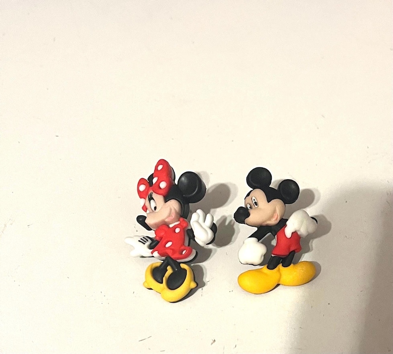 May include: Two cartoon figurines of Mickey and Minnie Mouse. Minnie is wearing a red dress with white polka dots and a red bow. Mickey is wearing red shorts and white gloves. Both have yellow shoes and black ears.