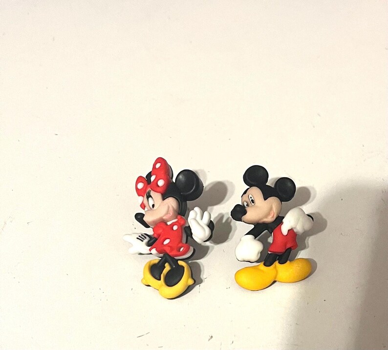 May include: Two cartoon character figurines, Minnie Mouse and Mickey Mouse, stand side-by-side. Minnie wears a red dress with white polka dots and a matching bow. Mickey wears red shorts and yellow shoes. Both have black ears.