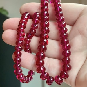 May include: A necklace of polished, deep red beads. The beads are round and evenly sized, strung together to form a necklace. The colour is a rich, vibrant red, with a glossy finish. The necklace is held in a hand.