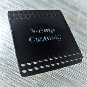 May include: A black square-shaped metal plate with a grid pattern around the edges and the text "V-Amp Customs" engraved in the center.