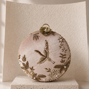 May include: A round, blush pink velvet clutch bag featuring intricate gold thread embroidery of a crane in flight, surrounded by floral motifs, small white pearls, and a single olive green fabric flower at the top with a gold metal ring handle.