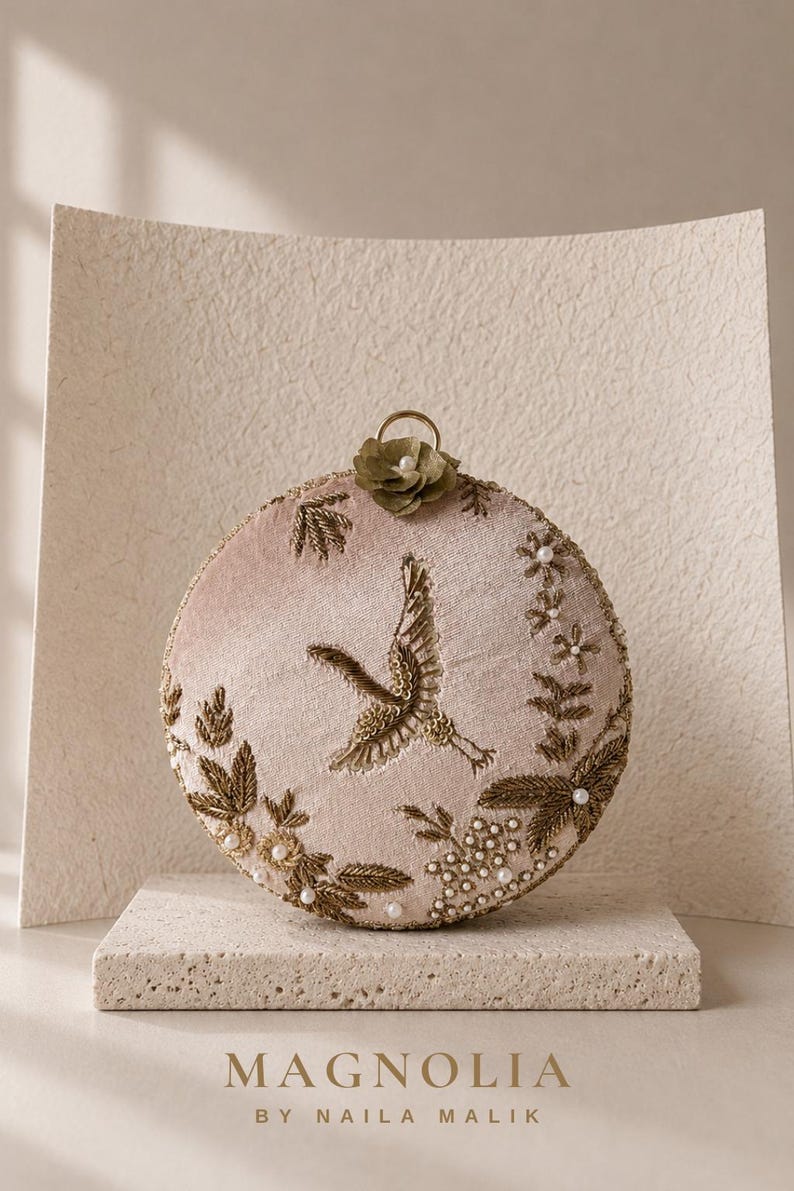 May include: A round, blush pink velvet clutch bag featuring intricate gold thread embroidery of a crane in flight, surrounded by floral motifs, small white pearls, and a single olive green fabric flower at the top with a gold metal ring handle.