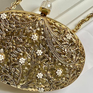 May include: A gold metal clutch bag with a floral design and white pearl accents. The bag has a chain strap and a clear crystal top.