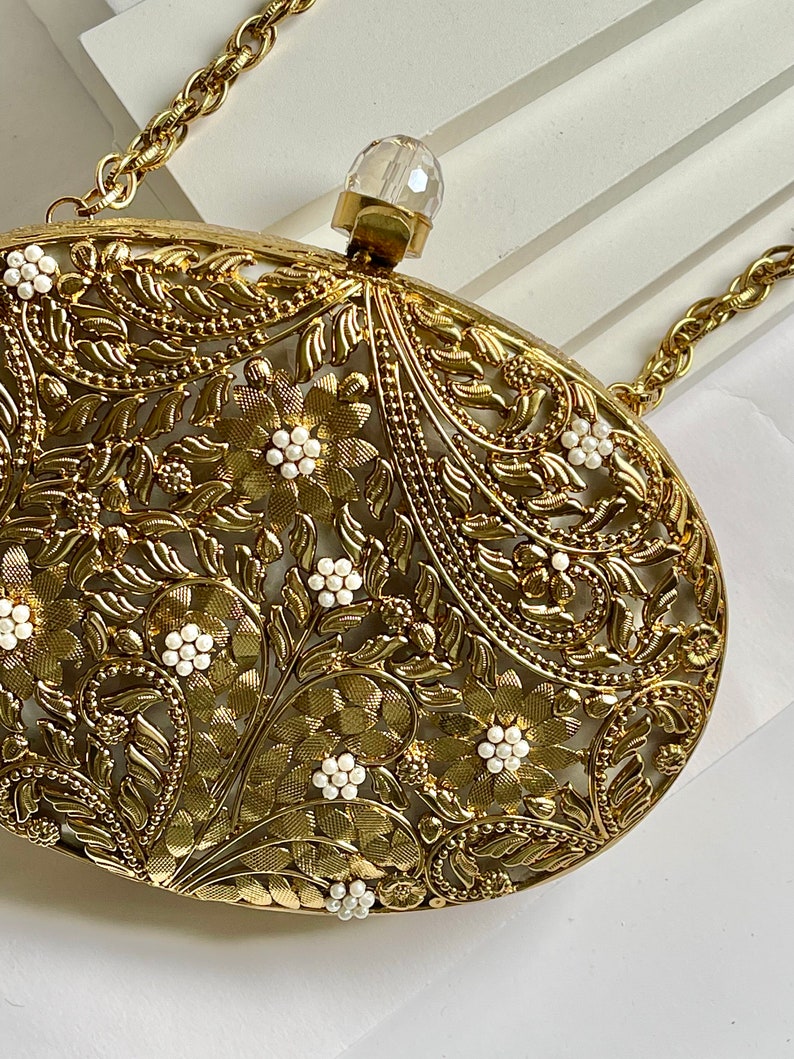 May include: A gold metal clutch bag with a floral design and white pearl accents. The bag has a chain strap and a clear crystal top.