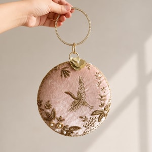 May include: A circular blush pink velvet handbag featuring gold metallic thread embroidery of a crane and floral motifs, accented with small white pearls. The bag has a round gold metal handle encrusted with clear crystals and a small flower charm.