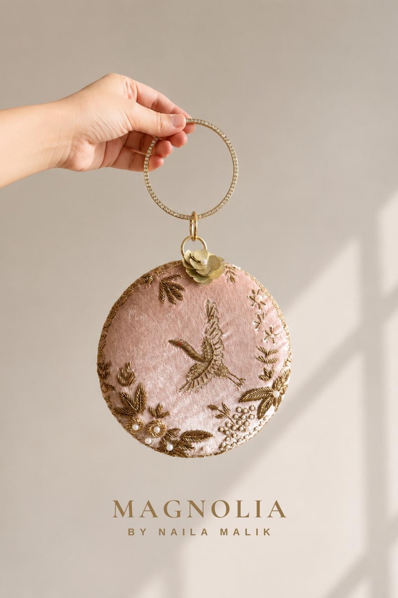 May include: A circular blush pink velvet handbag featuring gold metallic thread embroidery of a crane and floral motifs, accented with small white pearls. The bag has a round gold metal handle encrusted with clear crystals and a small flower charm.