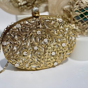 May include: A gold clutch bag with a white floral design and pearl accents. The bag has a chain strap.