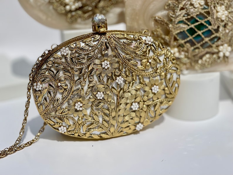 May include: A gold clutch bag with a white floral design and pearl accents. The bag has a chain strap.