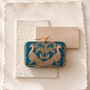 May include: Teal clutch with gold embroidery featuring two peacocks and a tree design. The clutch has a gold bird clasp and is labeled "MAGNOLIA BY NAILA MALIK".