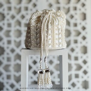 May include: Ivory beaded pouch with a drawstring closure. The bag is decorated with pearls and silver accents, and has a tassel with clear crystal accents. The bag is displayed on a white stand against a white patterned background.