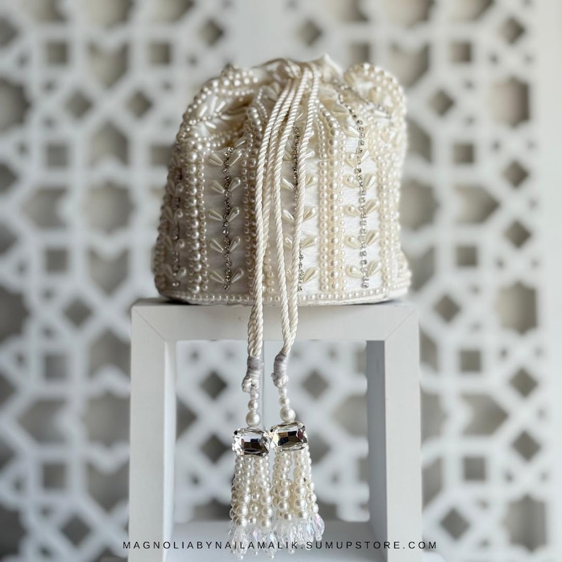 May include: Ivory beaded pouch with a drawstring closure. The bag is decorated with pearls and silver accents, and has a tassel with clear crystal accents. The bag is displayed on a white stand against a white patterned background.