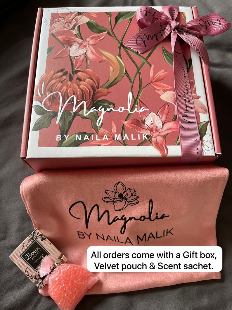 May include: A pink gift box with a floral design and a pink ribbon. The box says "Magnolia by Naila Malik". The box is sitting on a pink velvet pouch with the same logo. The pouch is next to a small pink and white sachet.