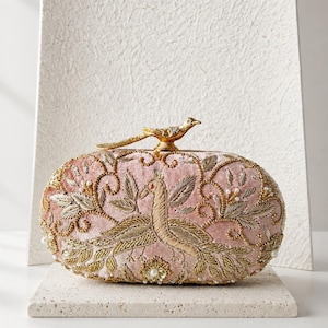 May include: Pink embroidered clutch with gold detailing and a bird clasp. The clutch features an embroidered peacock design and is embellished with pearls. The text "MAGNOLIA BY NAILA MALIK" is displayed below the clutch.