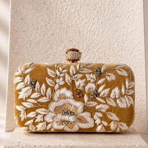 Könnte beinhalten: Senffarbene Samt-Clutch mit aufwendiger weißer Blumenstickerei und einem Schmuckverschluss. Die rechteckige Tasche hat ein großes Blumenmuster und trägt die Aufschrift "MAGNOLIA" von Naila Malik.