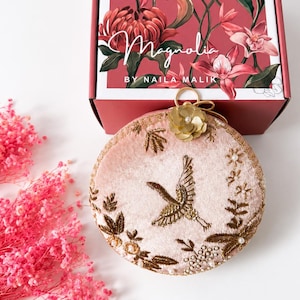 May include: A round, pale pink velvet clutch bag featuring gold thread embroidery of a crane and floral motifs, accented with a gold flower embellishment. It is displayed in front of a box labeled Magnolia by Naila Malik with floral graphics.