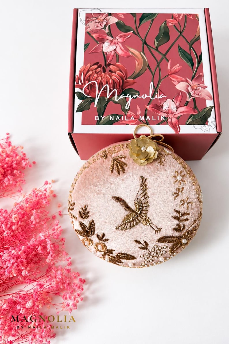 May include: A round, pale pink velvet clutch bag featuring gold thread embroidery of a crane and floral motifs, accented with a gold flower embellishment. It is displayed in front of a box labeled Magnolia by Naila Malik with floral graphics.
