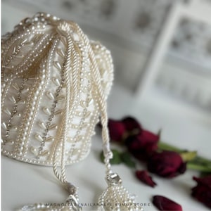 May include: Cream-coloured beaded drawstring bag with pearl and rhinestone accents. The bag has a tassel and a drawstring closure. The bag is decorated with pearls and rhinestones. The text "MAGNOLIABYNAILAMALIK.SUMUPSTORE." is at the bottom.