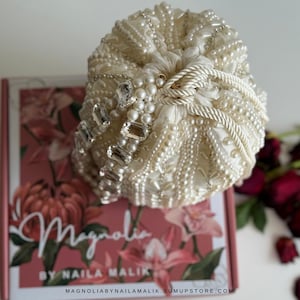 May include: Cream-coloured, beaded turban-style head covering with crystal and pearl detailing. The head covering is placed on a box with the word "Magnolia" and floral designs.