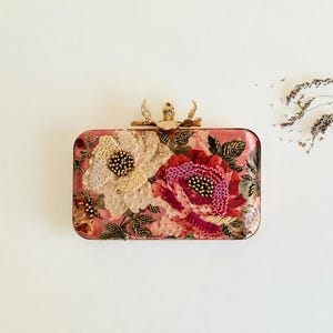 May include: A rectangular clutch bag featuring a pink base with intricate floral embroidery in shades of cream, red, and magenta. The surface is embellished with beads and sequins, and the top closure is a gold-toned metal flower-shaped clasp.