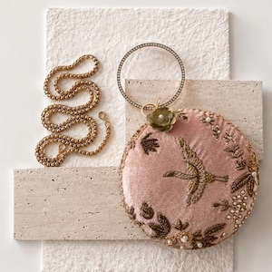 May include: A round, dusty pink velvet clutch bag featuring gold-toned embroidered bird and floral motifs, accented with small white pearls and a circular crystal-encrusted handle. A gold-toned chain strap is included. Text reads: MAGNOLIA BY NAILA MALIK.