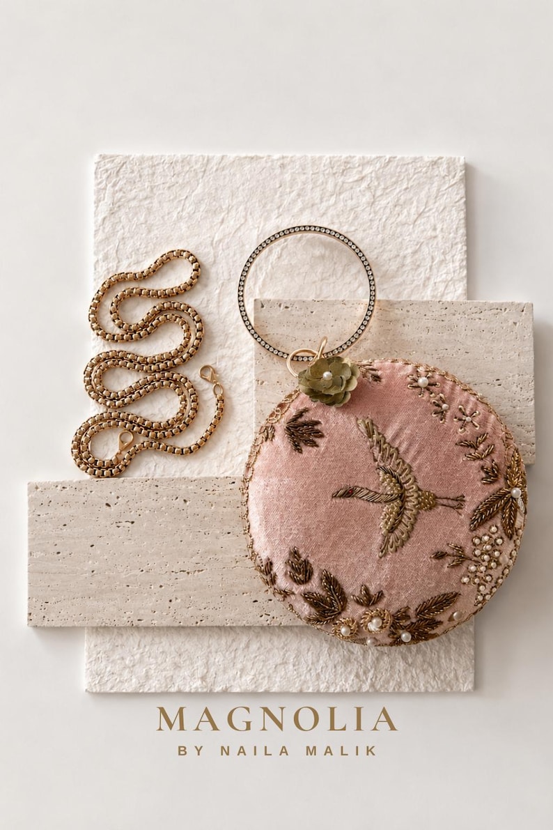 May include: A round, dusty pink velvet clutch bag featuring gold-toned embroidered bird and floral motifs, accented with small white pearls and a circular crystal-encrusted handle. A gold-toned chain strap is included. Text reads: MAGNOLIA BY NAILA MALIK.