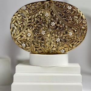 May include: A gold metal clutch bag with an intricate floral design and white pearl accents. The bag is on a white pedestal.