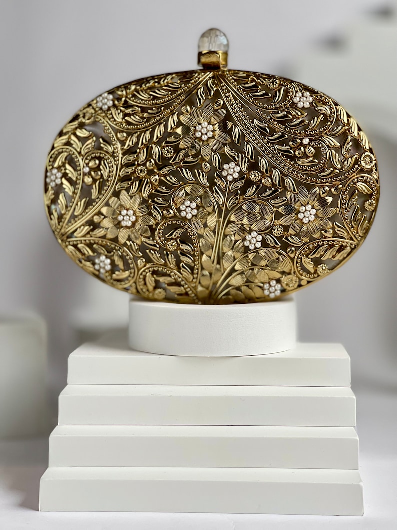 May include: A gold metal clutch bag with an intricate floral design and white pearl accents. The bag is on a white pedestal.