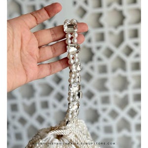 May include: A white beaded handbag with a handle made of small pearls and large, rectangular, clear crystals. The bag's bottom edge is visible, with the text "MAGNOLIABYNAILAMALIK.SUMUPSTORE.COM".