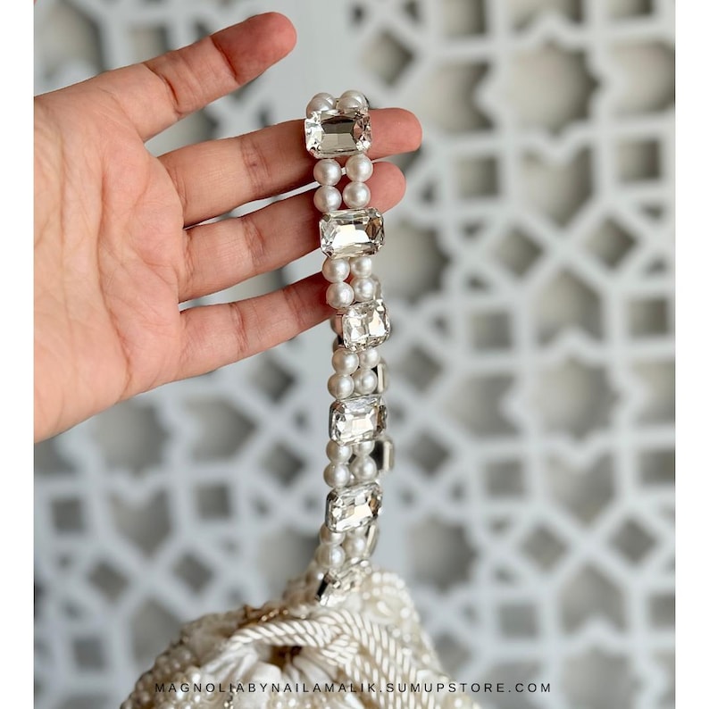 May include: A white beaded handbag with a handle made of small pearls and large, rectangular, clear crystals. The bag's bottom edge is visible, with the text "MAGNOLIABYNAILAMALIK.SUMUPSTORE.COM".