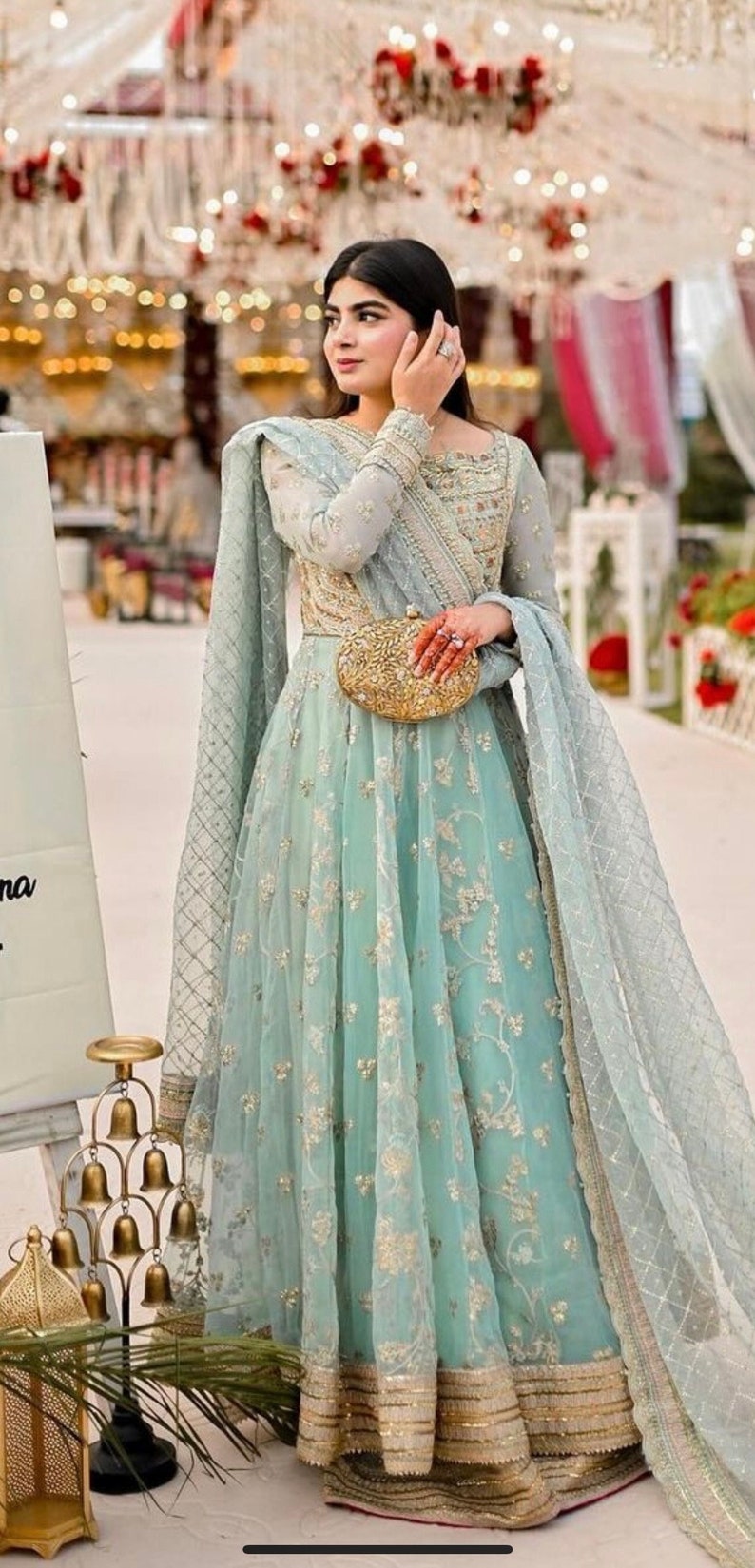 May include: A woman wearing a light blue, embroidered dress with a gold clutch. The dress has a long, flowing skirt and a fitted bodice. The woman is standing in front of a white backdrop with gold decorations.