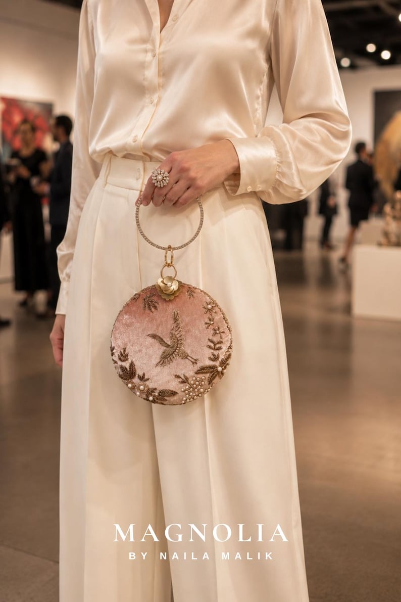 May include: A round, blush pink velvet handbag featuring gold-toned embroidery of a crane and floral motifs, accented with small white pearls. The bag has a circular metal handle encrusted with clear crystals and a gold-toned flower clasp.