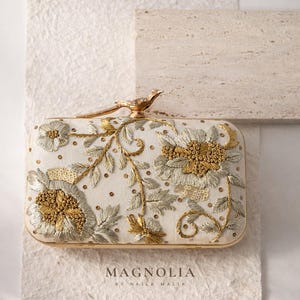 May include: Ivory rectangular clutch bag with gold and silver floral embroidery, sequins, and a gold bird clasp. The bag features the text "MAGNOLIA BY NAILA MALIK" at the bottom. A decorative accessory.