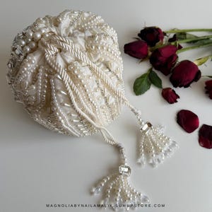 May include: Cream-coloured drawstring bag adorned with pearls and crystals. The bag has a gathered design, rope-style drawstrings, and beaded tassels. The text "MAGNOLIABYNAILAMALIK.SUMUPSTORE.COM" is visible. Dried red roses are in the background.
