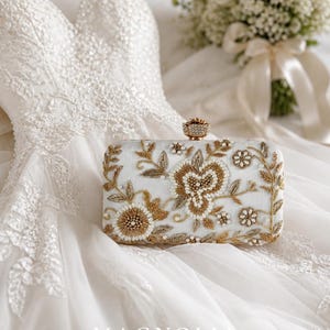 May include: A white rectangular clutch with intricate gold floral embroidery and pearl accents. The clutch features a jeweled clasp and is placed on a white wedding dress. The text "MAGNOLIA BY NAILA MALIK" is visible.