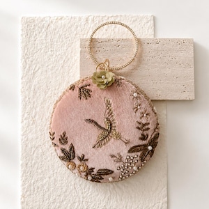 May include: A circular dusty pink velvet handbag featuring intricate gold thread embroidery of a crane in flight, surrounded by floral motifs, small white beads, and a single olive green fabric flower. It has a thin, circular gold metal handle.