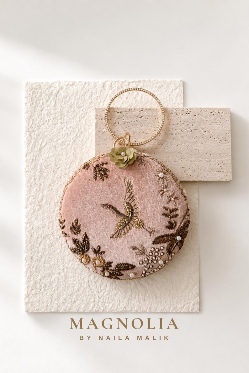 May include: A circular dusty pink velvet handbag featuring intricate gold thread embroidery of a crane in flight, surrounded by floral motifs, small white beads, and a single olive green fabric flower. It has a thin, circular gold metal handle.