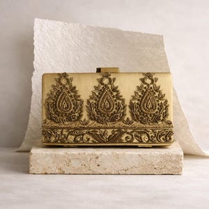 Könnte beinhalten: Eine rechteckige goldene Clutch mit aufwendiger, erhabener Stickerei. Das Design zeigt drei zentrale tropfenförmige Motive und detaillierte florale Muster. Die Clutch liegt auf einem Steinblock vor neutralem Hintergrund.