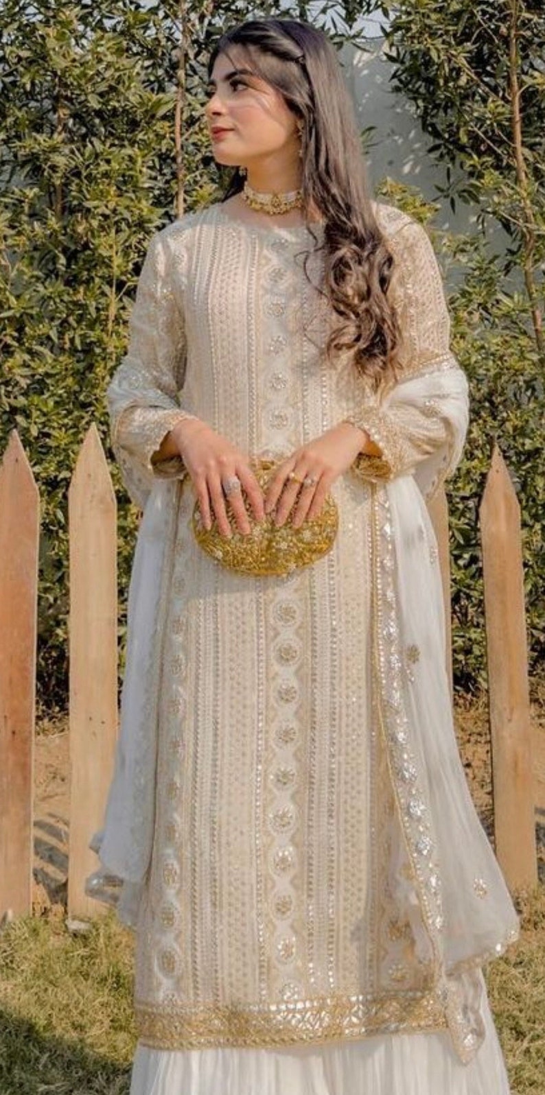 May include: A white embroidered dress with a gold clutch. The dress has a long sleeve and a white dupatta with gold embroidery. The dress is worn by a person standing in front of a wooden fence.