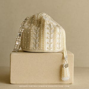 May include: Cream-coloured beaded pouch with a drawstring and tassel. The pouch is adorned with pearls and rectangular crystals on the strap. The design features vertical pearl rows and leaf-shaped details. The pouch rests on a beige block.