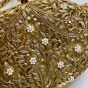 May include: A gold metal clutch bag with a floral design and white pearl accents. The bag has a chain strap.